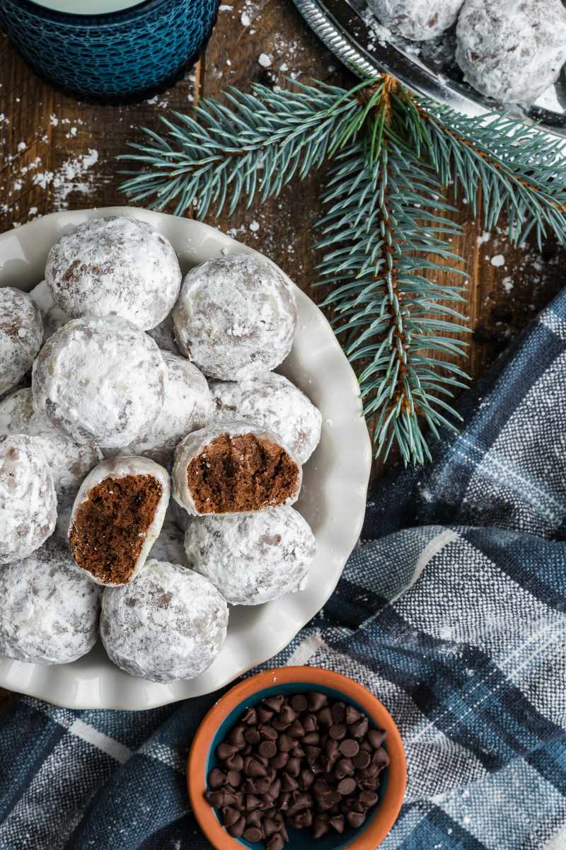 Top view of chocolate snowball cookies on a white plate on a wood table with chocolate chips and evergreen branches around.
