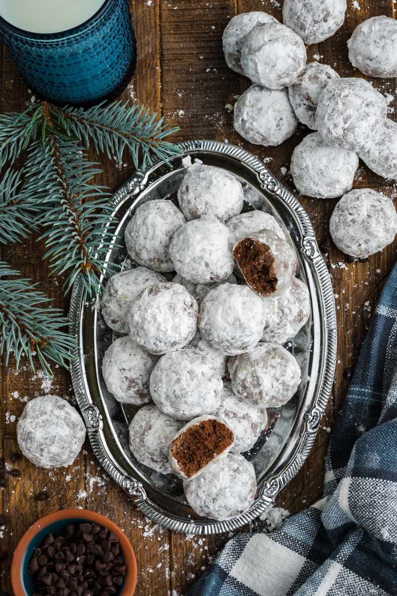 Overhead of chocolate snowballs on an oval silver platter with evergreen branches, chocolate chips, and more cookies around it.