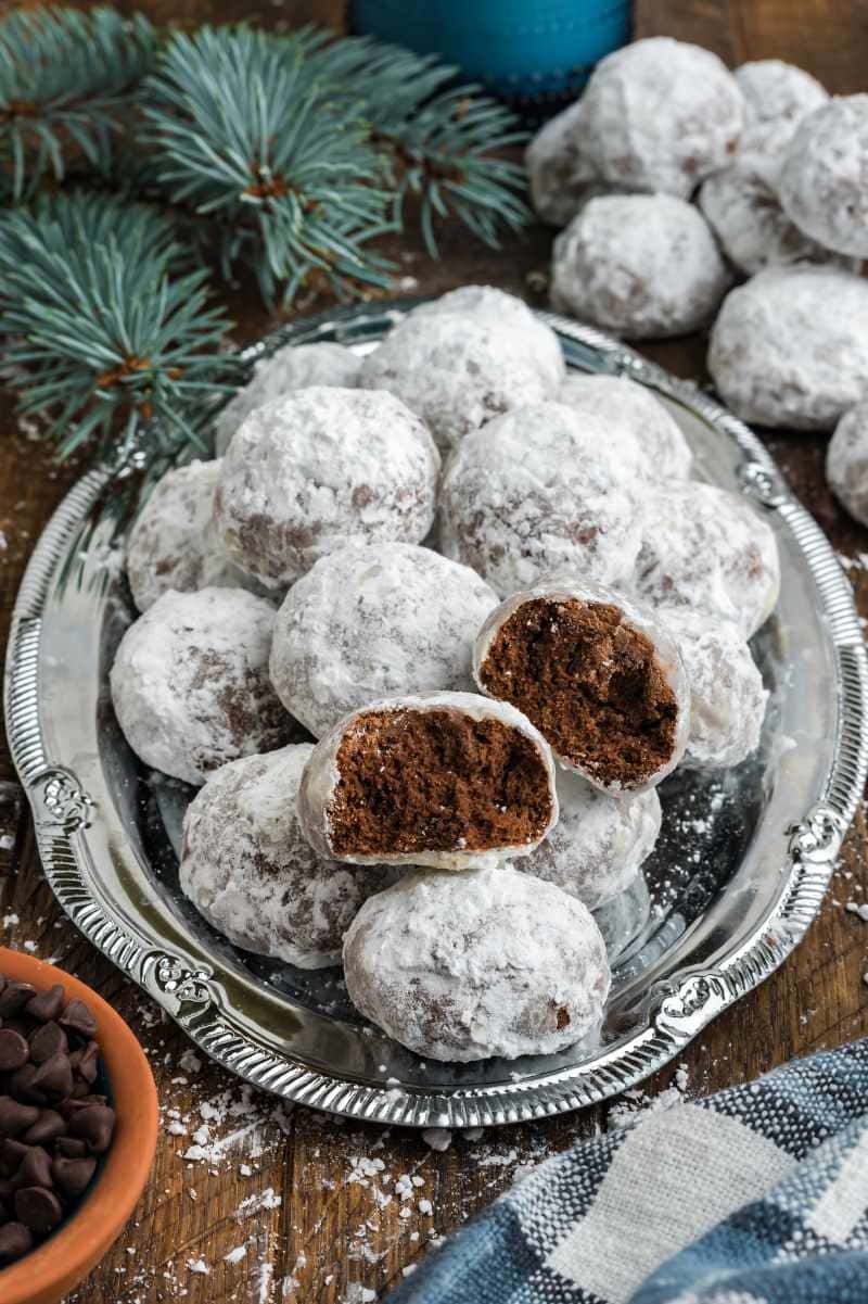 A platter of Christmas cookies rolled in powdered sugar rests on a holiday table, one cookie split open to show the chocolate middle.