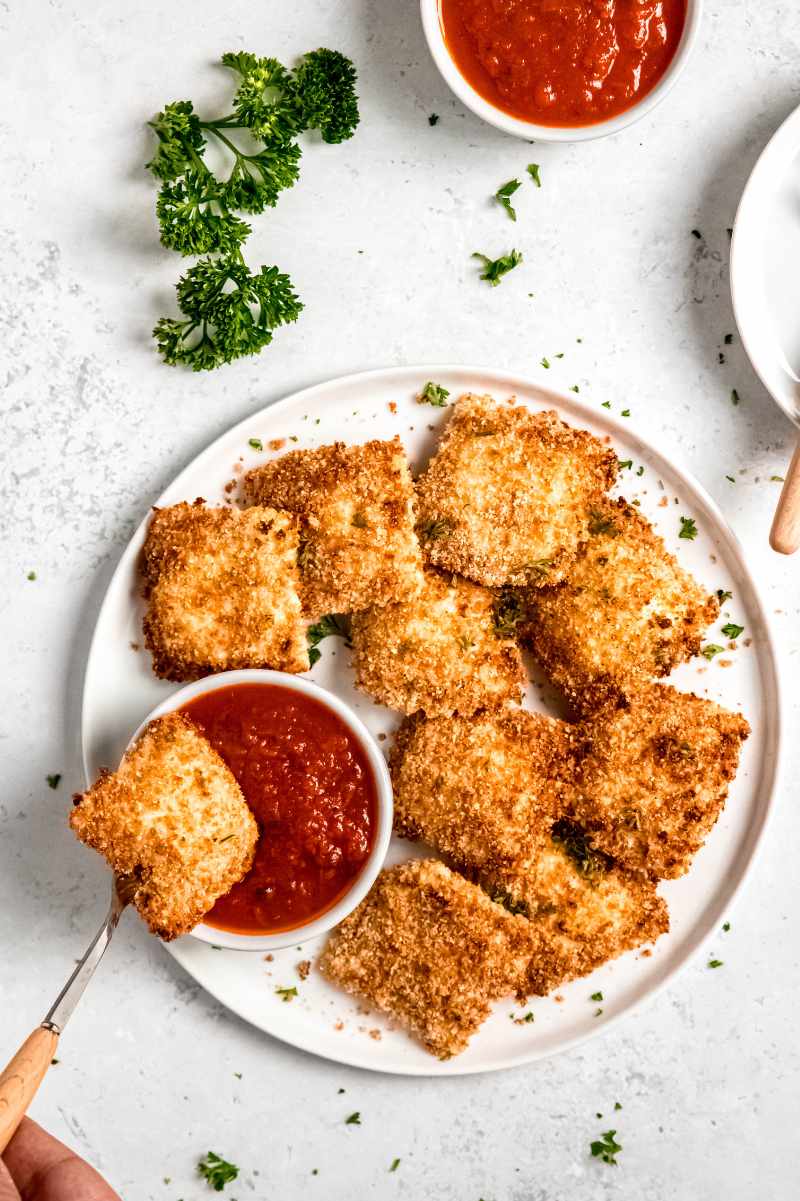 A fork dips a toasted ravioli into a small bowl of marinara sauce on a large white serving platter filled with raviolis.