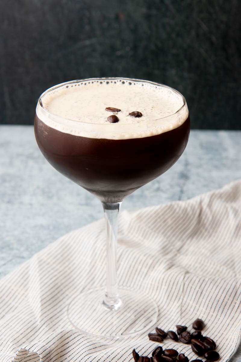 A coupe glass filled with an espresso martini stands on a kitchen linen with additional beans at the foot of the glass.