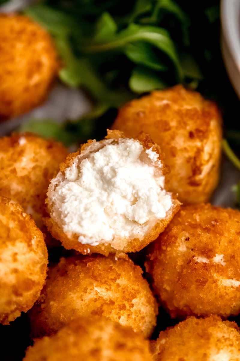 Half a fried goat cheese ball rests atop other crispy, golden goat cheese balls showing the creamy center.