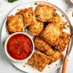 Toasted ravioli served on a white plate with a small bowl of marinara sauce and serving forks nearby.