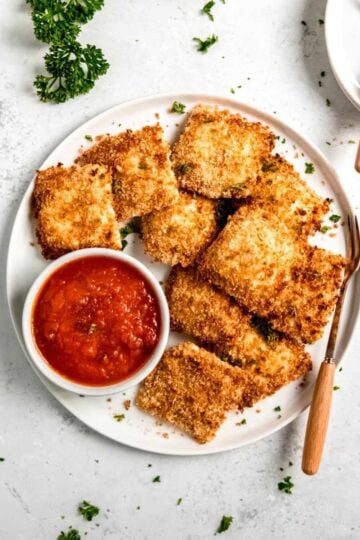 Toasted ravioli served on a white plate with a small bowl of marinara sauce and serving forks nearby.