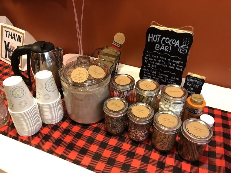 A hot cocoa bar sits on a red buffalo check covered table in a teacher's lounge at a school.
