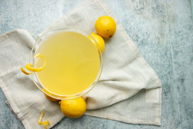 Top view of a limoncello cocktail on a cloth napkin with lemons around it on a grey counter.