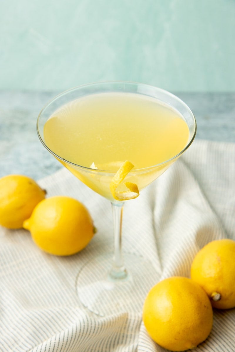 A limoncello martini stands on a striped kitchen linen on a grey counter with four whole lemons nearby.