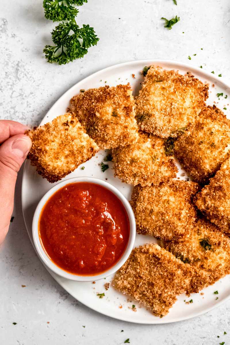 Two fingers grab a ravioli appetizer from a plate of raviolis and marinara sauce.