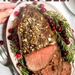 Two hands set a platter of roast beef on a light table with a large serving fork alongside. A text overlay reads, "Holiday Roast Beef."