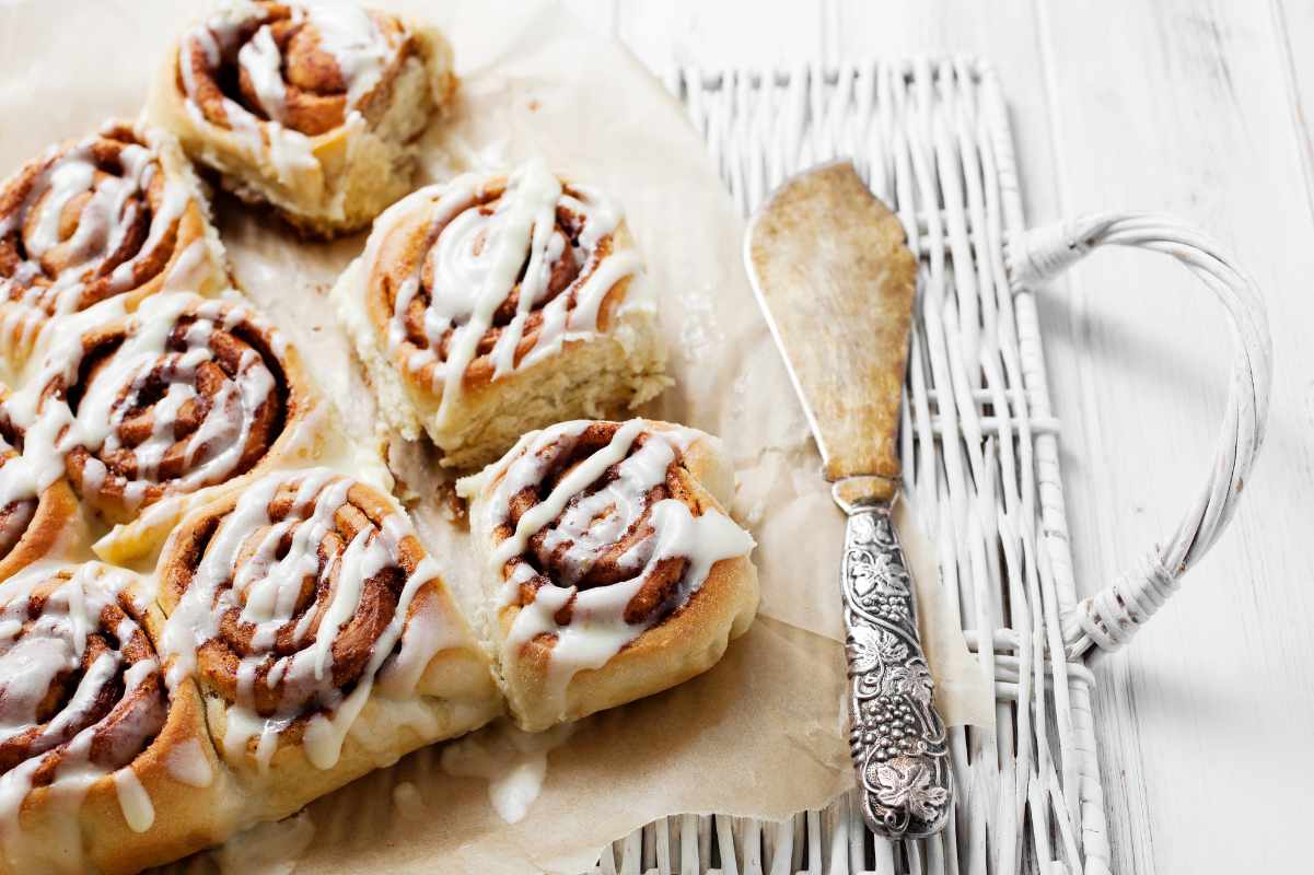 A white wicker serving tray with frosted cinnamon buns on parchment and a decorative serving knife.