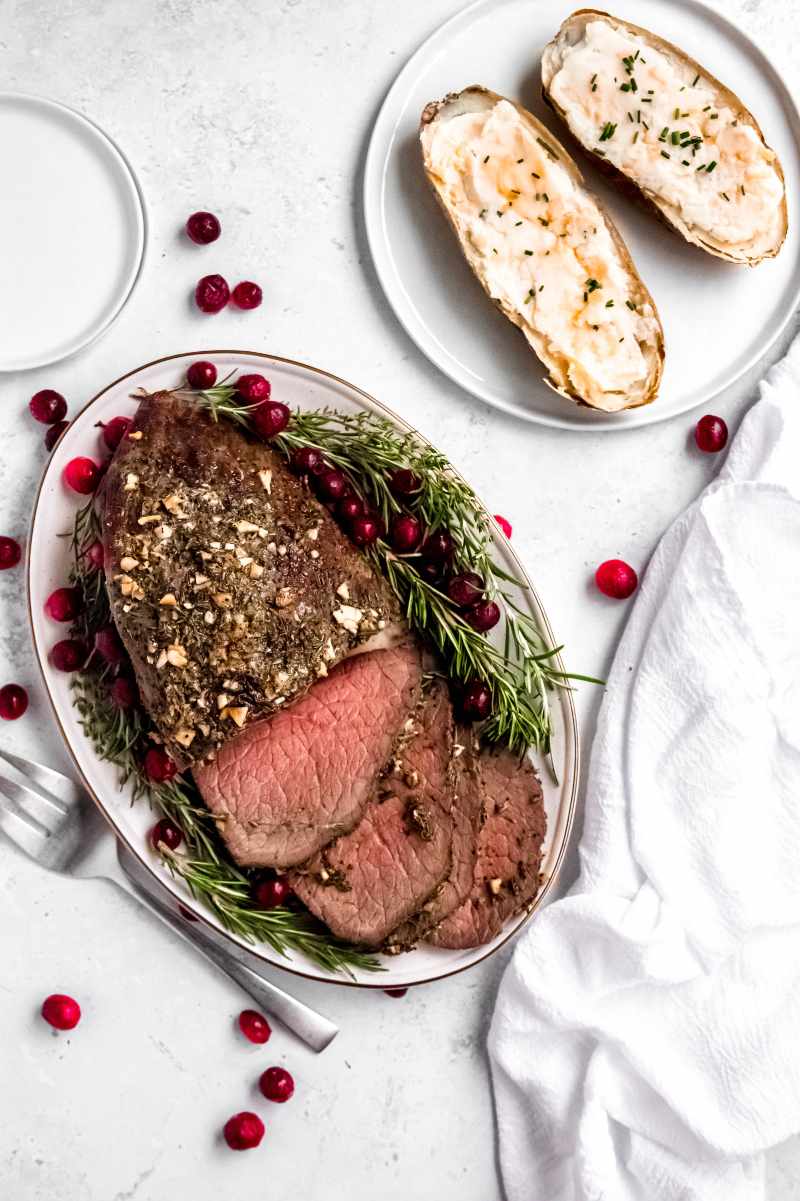 A tender beef dinner served on an oval platter with rosemary sprigs and fresh cranberries with a plate of twice baked potatoes alongside.