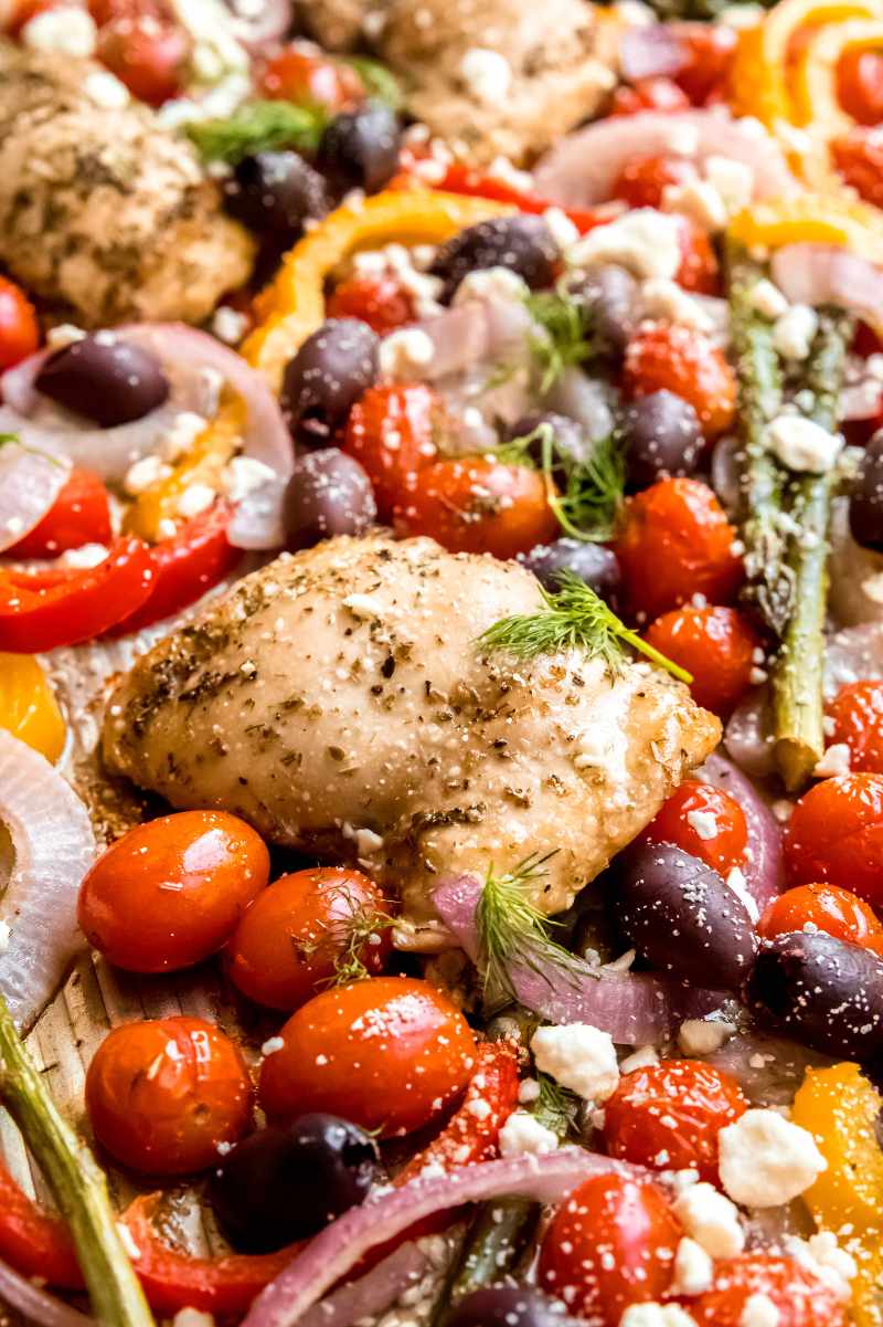 Front view of greek sheet pan chicken and vegetables such as grape tomatoes, onions, bell peppers, and asparagus.