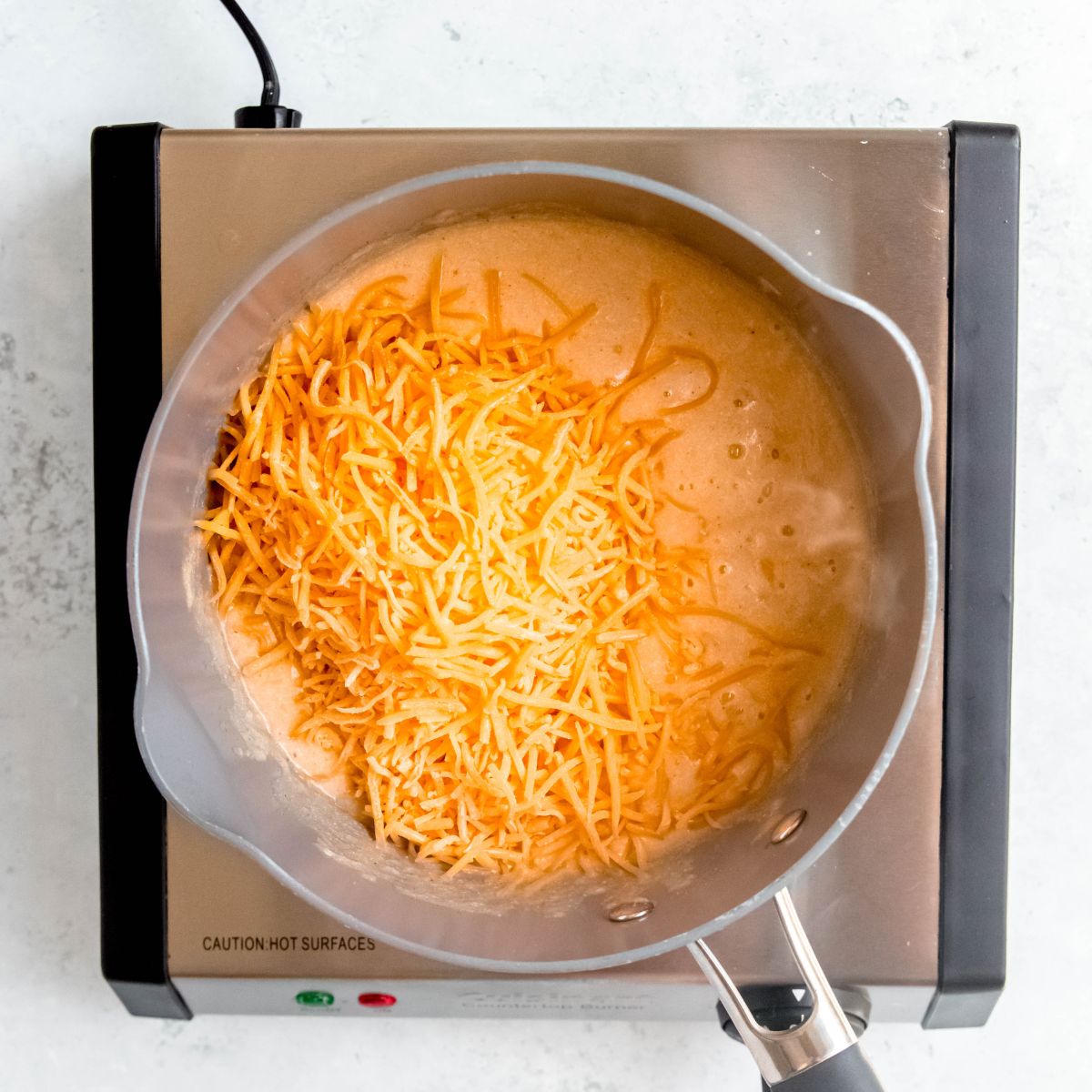Shredded cheddar cheese being added to a beer and flour mixture in a pot on a portable burner.