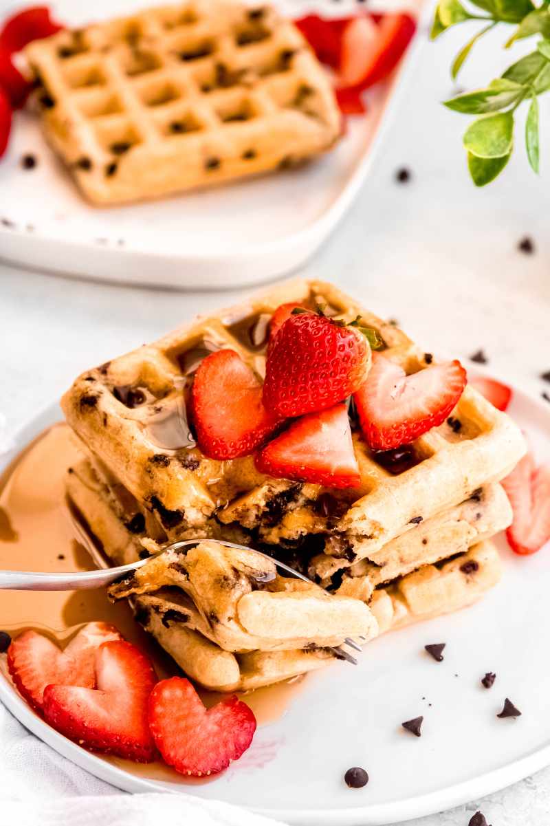A fork cuts into a stack of chocolate chip waffles topped with syrup and strawberries.