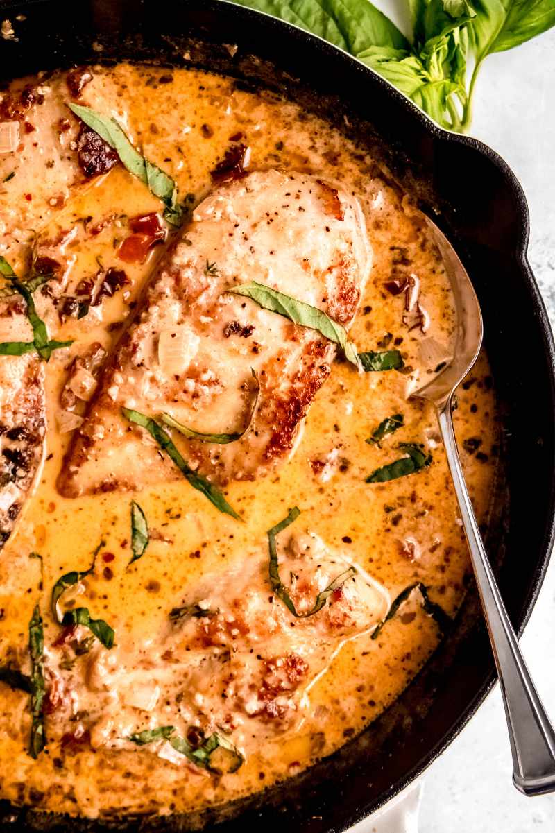 A spoon rests in a skillet filled with saucy chicken garnished with fresh basil.