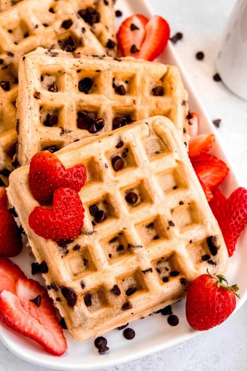 Homemade waffles studded with chocolate morsels on a white platter surrounded by cut strawberries.