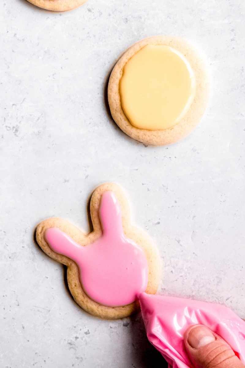 A hand squeezes pink frosting from a piping bag to decorate a bunny-shaped cookie.