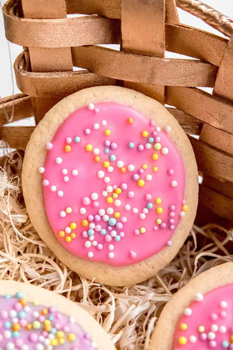And egg-shaped sugar cookie with pink frosting and pastel sprinkles stands against the side of a wooden Easter basket.