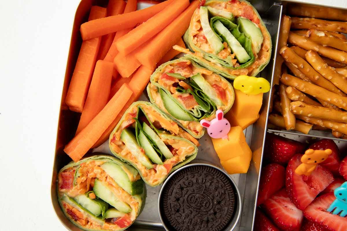 Veggie wrap bites with hummus packed into a school lunch with fruit, cheese, and snacks.