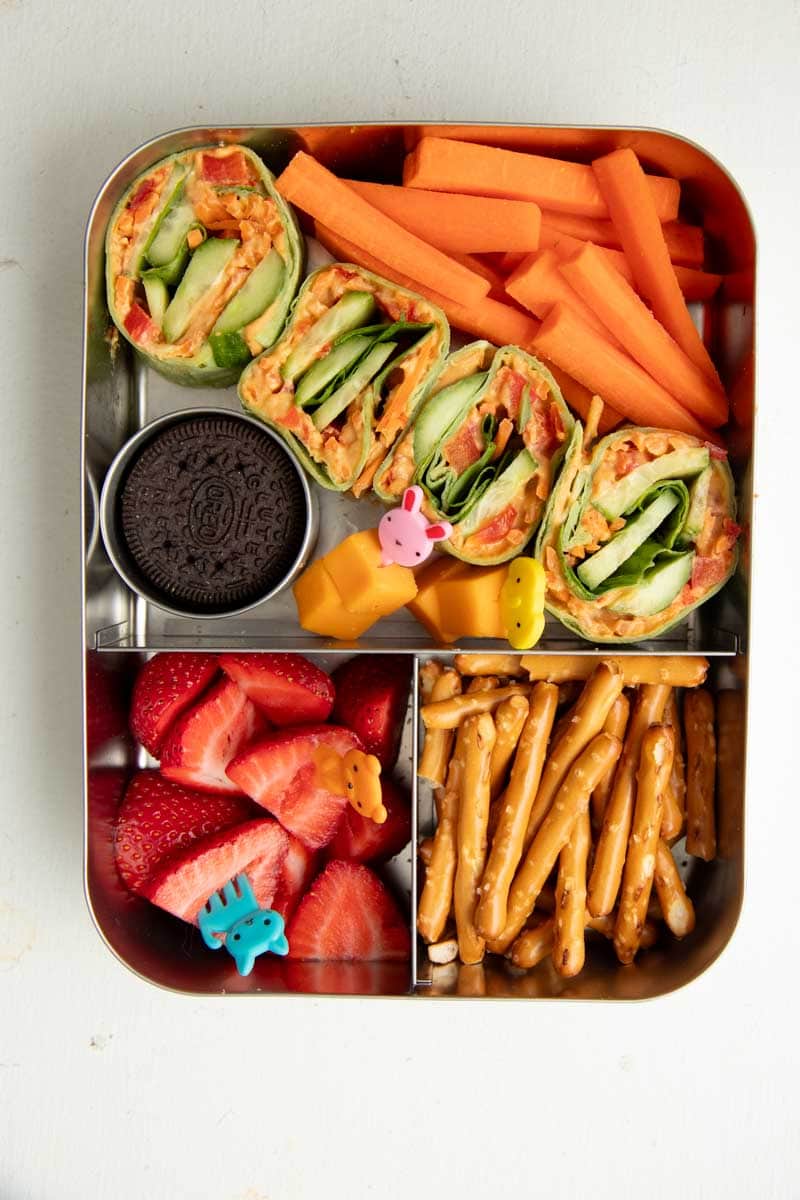Overhead of bento-style lunchbox pack with veggie pinwheels, carrot sticks, pretzel sticks, sliced strawberries, cheese cubes, and an oreo cookie.