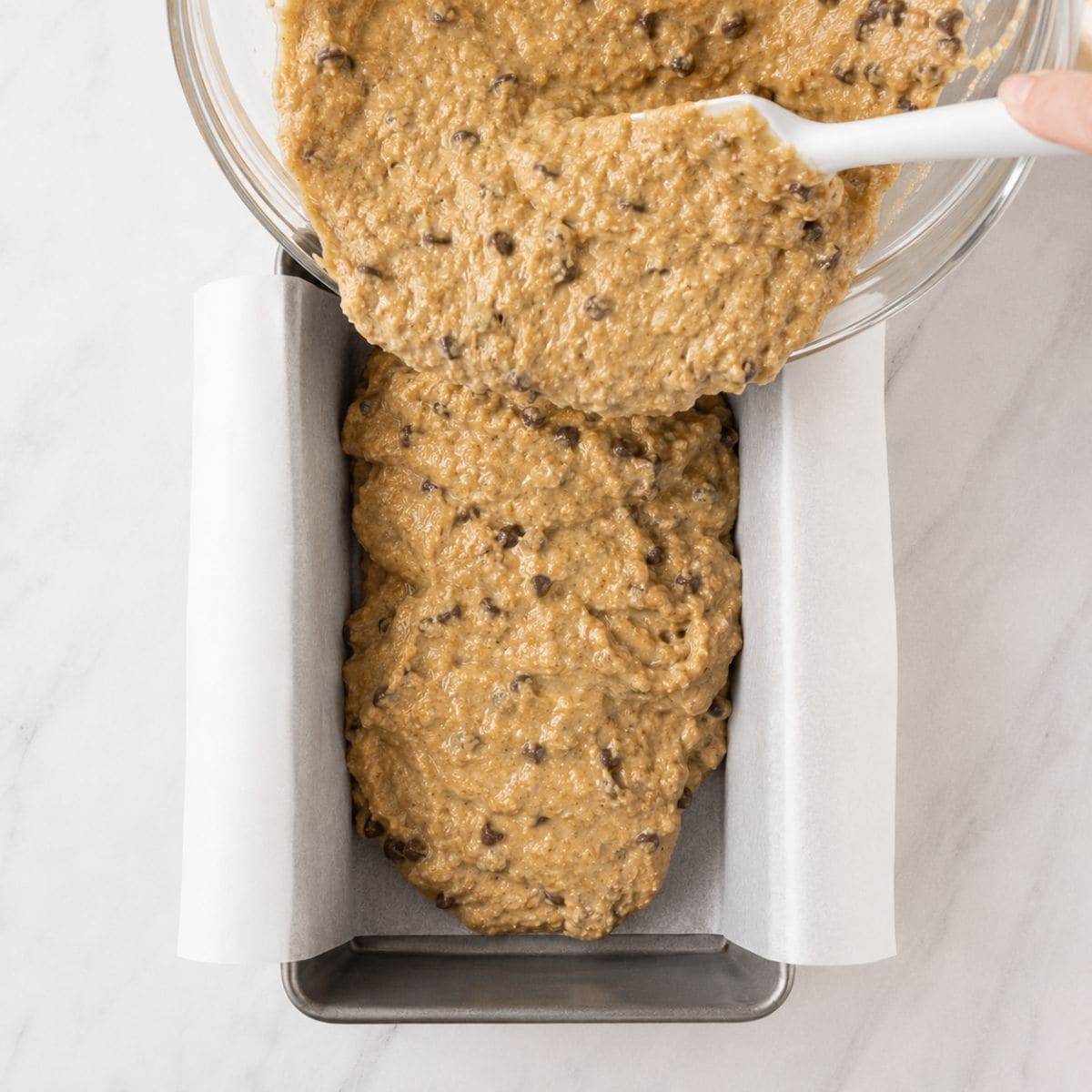 Chocolate chip banana bread batter being poured into a parchment-lined loaf pan.