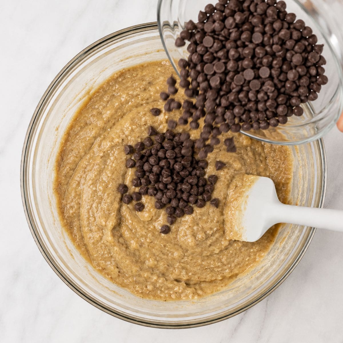 Mini chocolate chips being poured from a glass bowl into a bowl of banana bread batter.