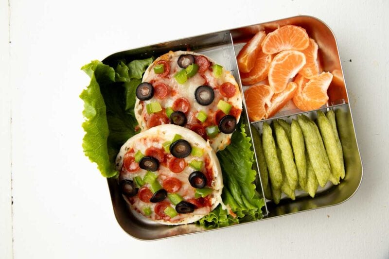 Overhead of English muffin pizzas in a bento-style lunch box with clementines and snap pea crisps.