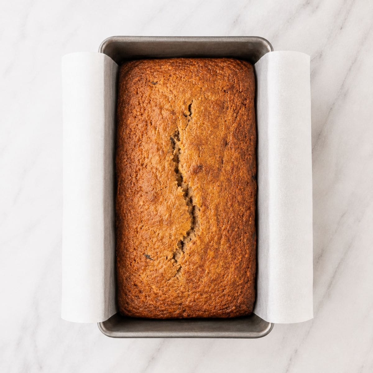 A golden baked loaf of banana bread cooling in its parchment-lined pan.