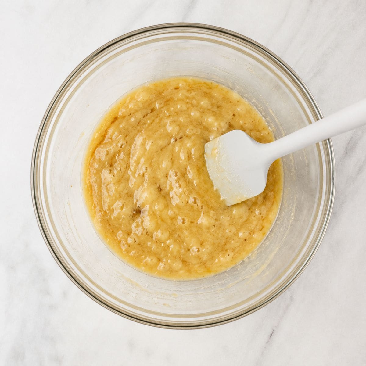 Mashed ripe bananas in a glass mixing bowl with a white rubber spatula.