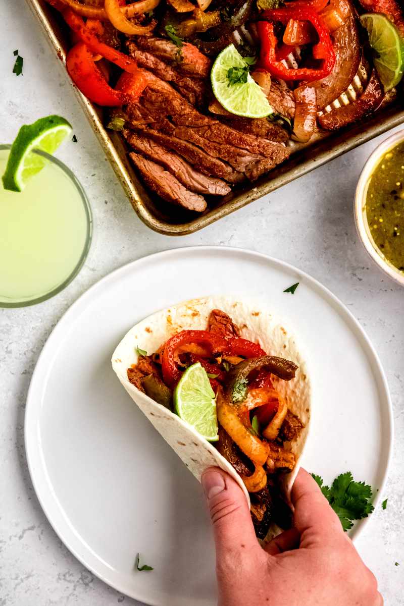 A hand picks up a soft tortilla stuffed with colorful fajitas meat and veggies from a white plate with salsa and a margarita nearby.