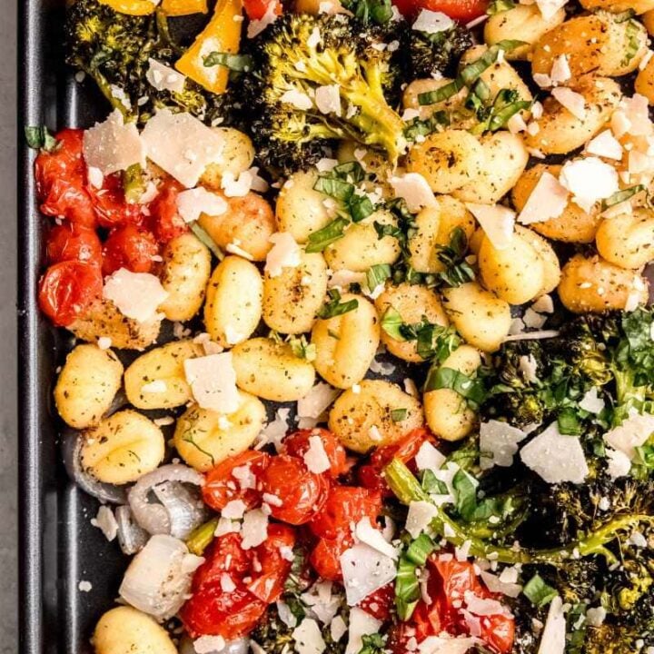 Top view of a dark sheet pan filled with baked gnocchi and vegetables topped with fresh basil and parmesan cheese.