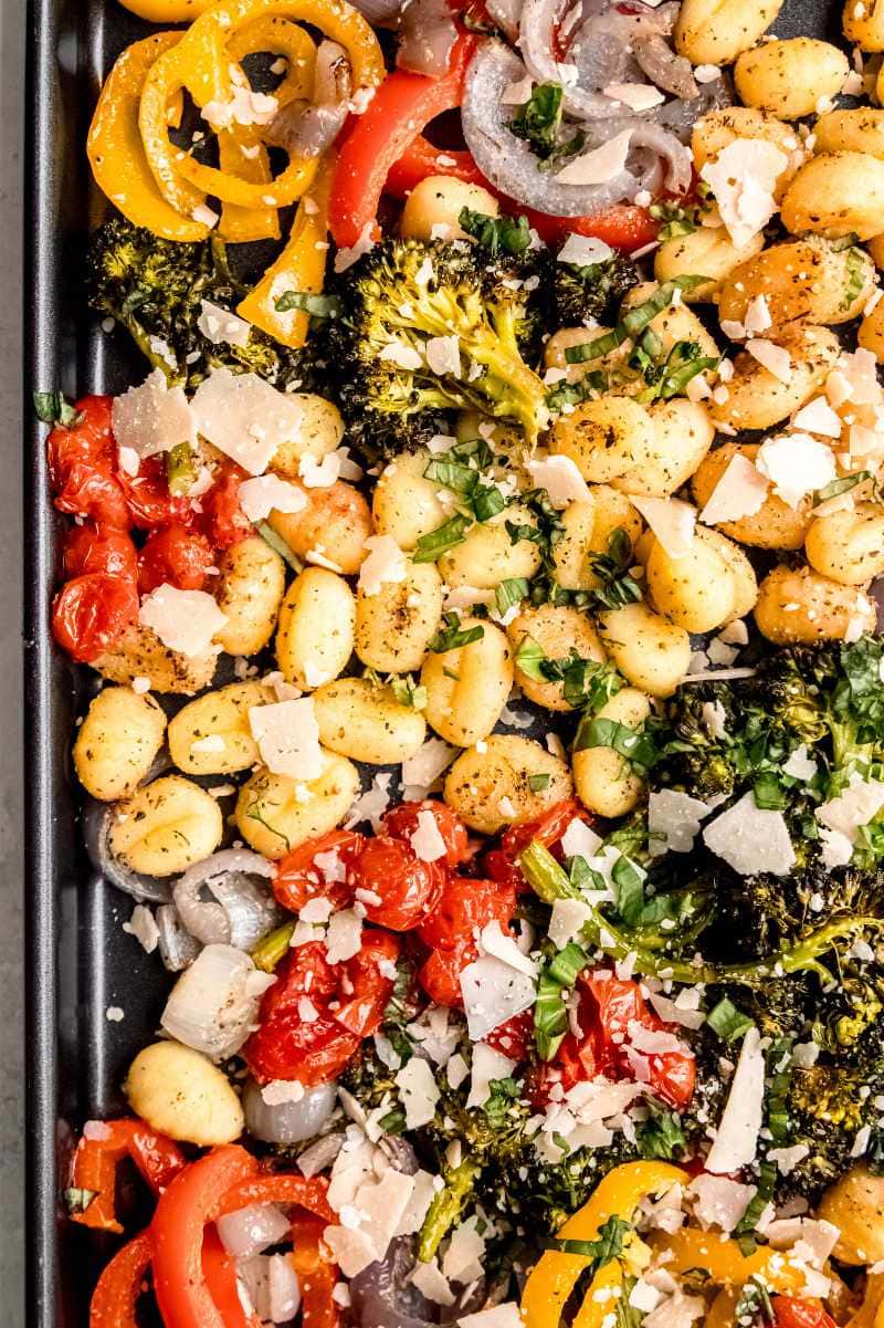 Dark sheet pan filled with baked gnocchi and vegetables topped with fresh basil and parmesan cheese.