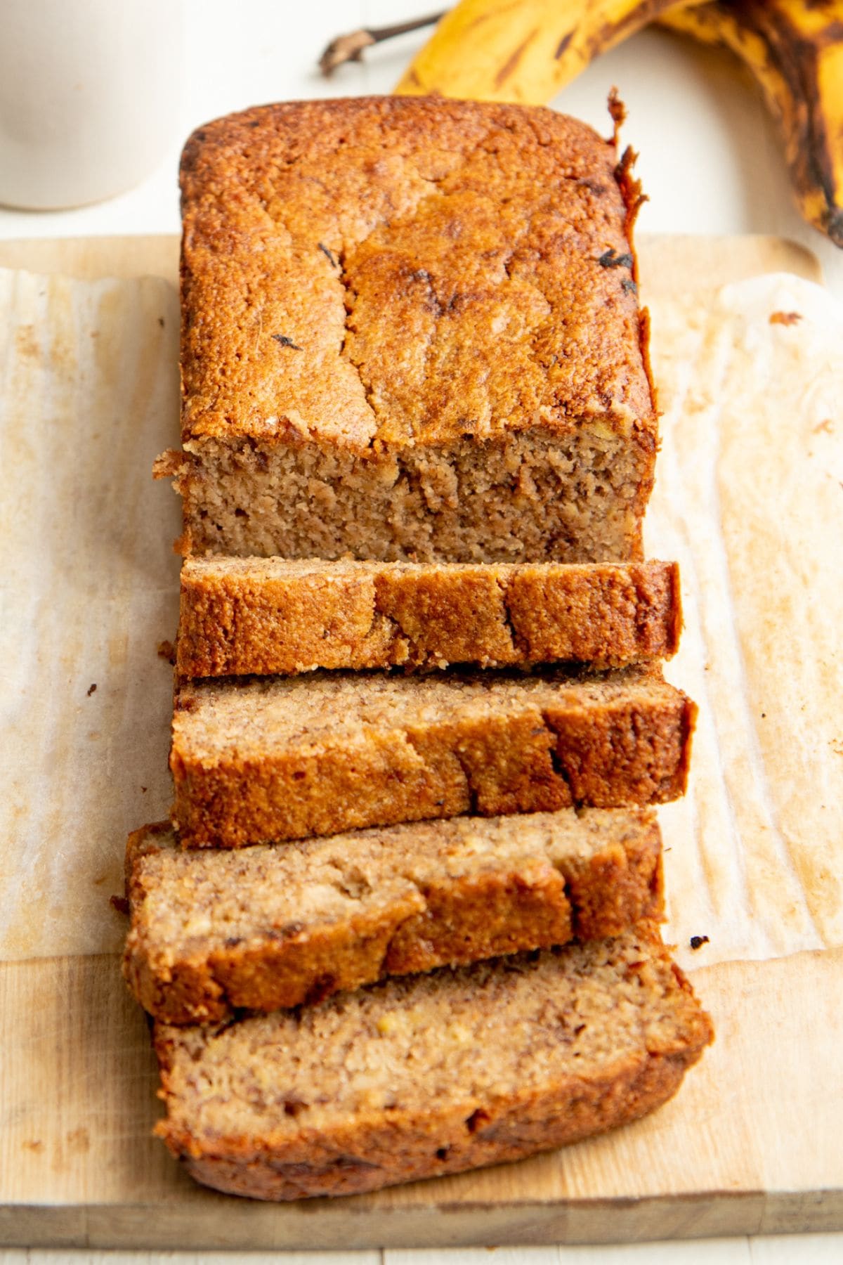 A loaf of banana bread sliced on a wooden board, with ripe bananas and a mug in the background.