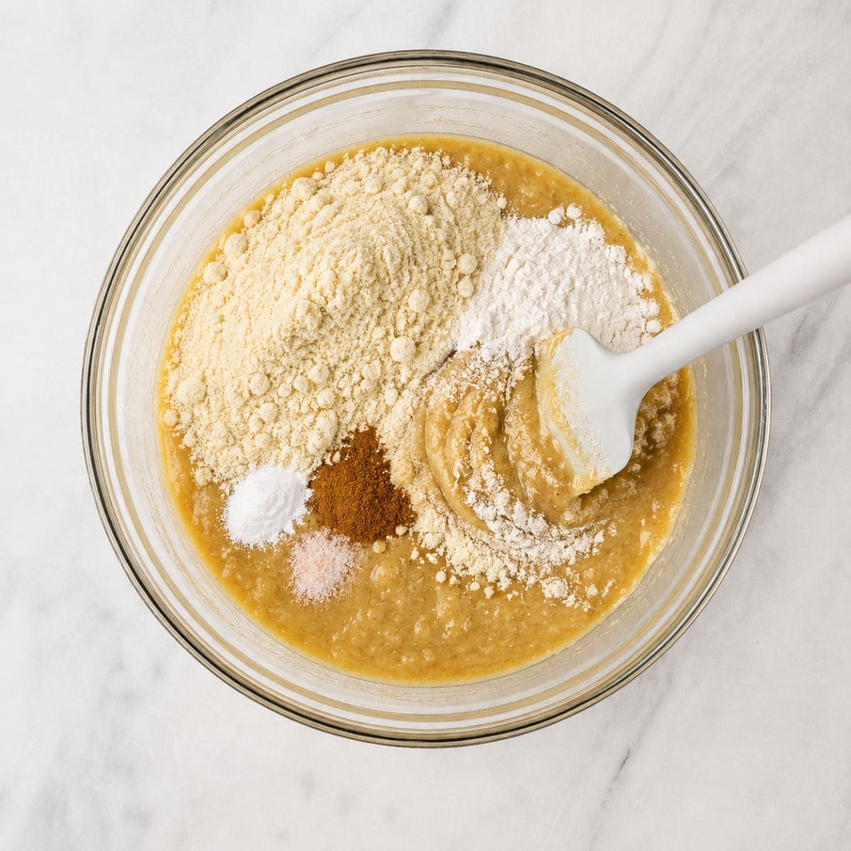 Almond flour, tapioca flour, cinnamon, baking soda, and salt added on top of a bowl of mashed banana batter.