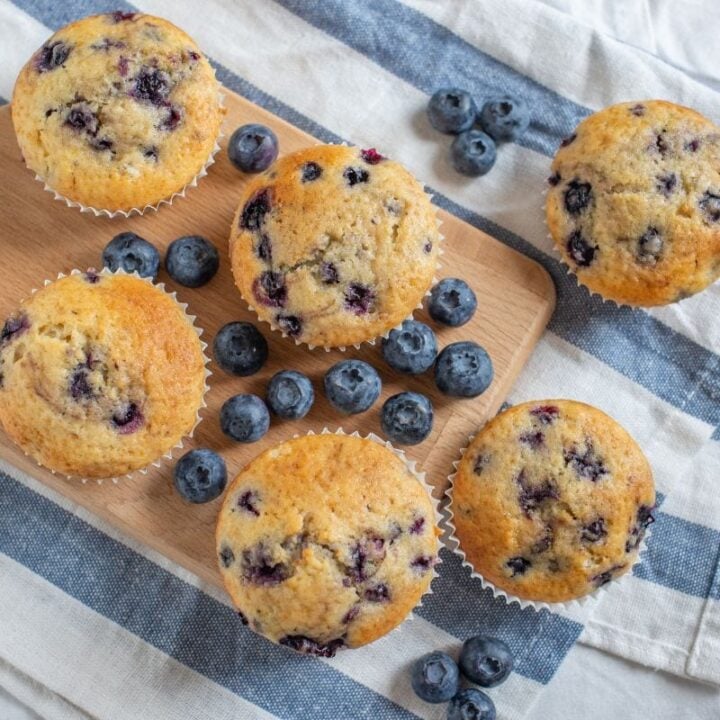 Top view of blueberry muffins on a wooden serving board with fresh berries and a blue and white striped kitchen linen.