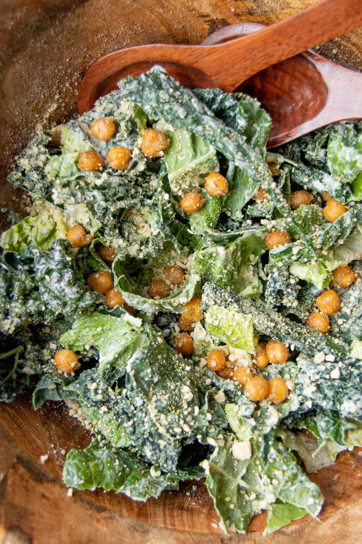 Dressed kale caesar salad showing the kale, roasted chickpeas, and grated parmesan mixed together, with a wooden spoon at the edge.