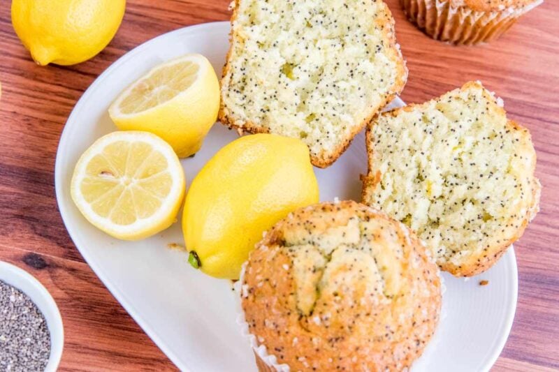 Top view of a lemon poppyseed muffin split on a white serving platter with additional lemons and another muffin.