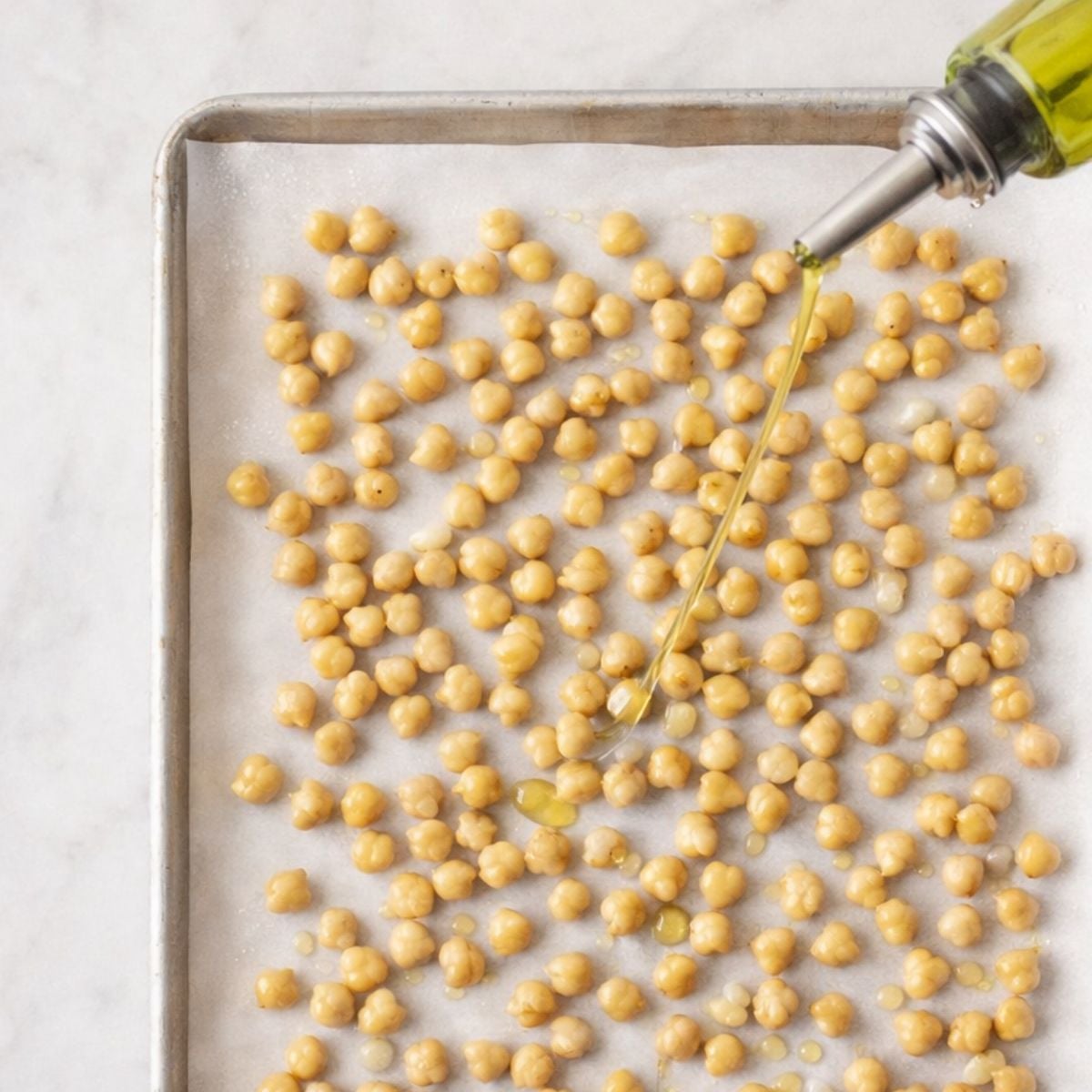Olive oil drizzled over chickpeas spread on a parchment-lined baking sheet before roasting them into croutons.