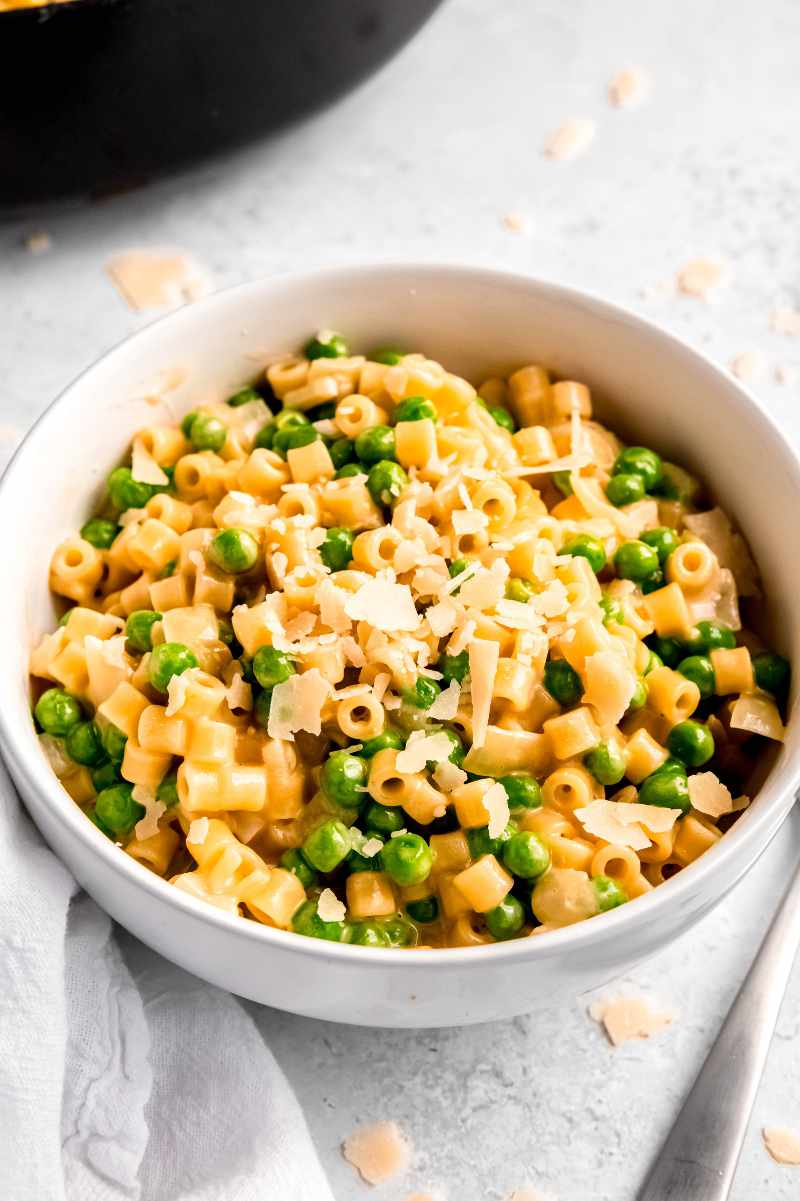 Creamy ditalini pasta with peas, garnished with shaved parmesan in a white bowl on a light counter.
