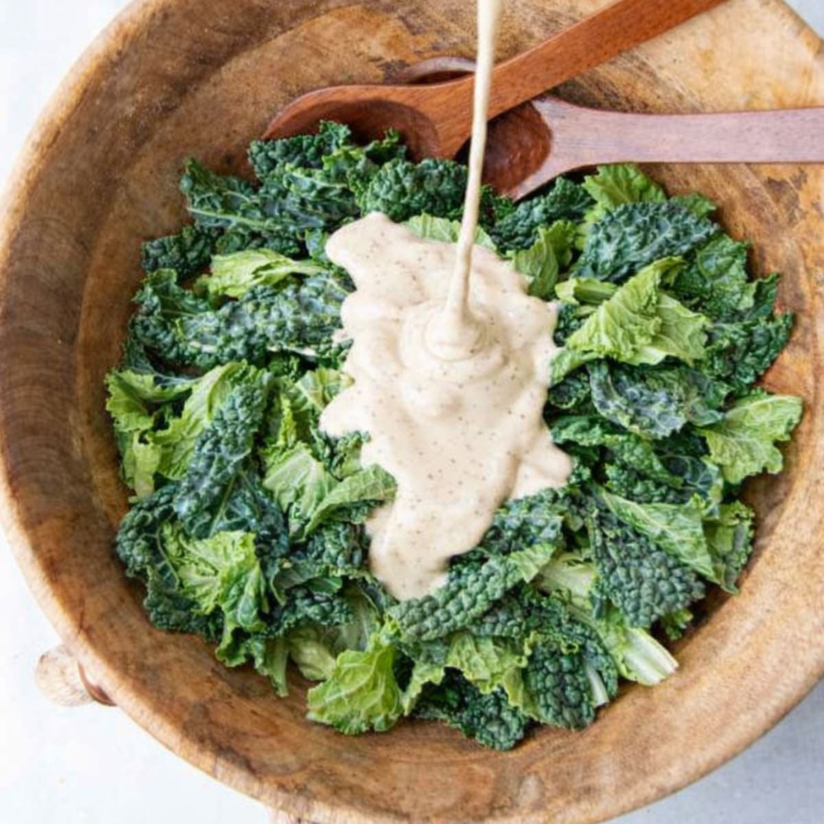 Creamy caesar dressing poured over a bowl of raw kale and romaine before tossing.