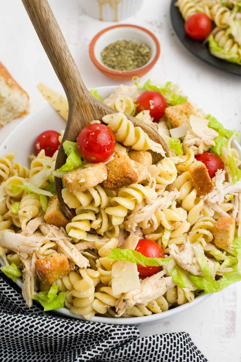 A wooden spoon scoops into a serving bowl of pasta salad.