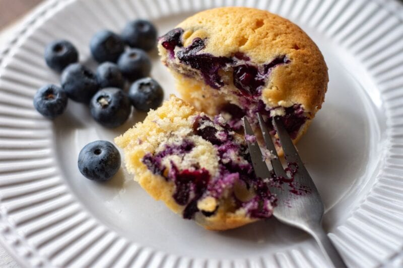 A dessert fork splits a blueberry muffin on a white plate with fresh berries beside it.