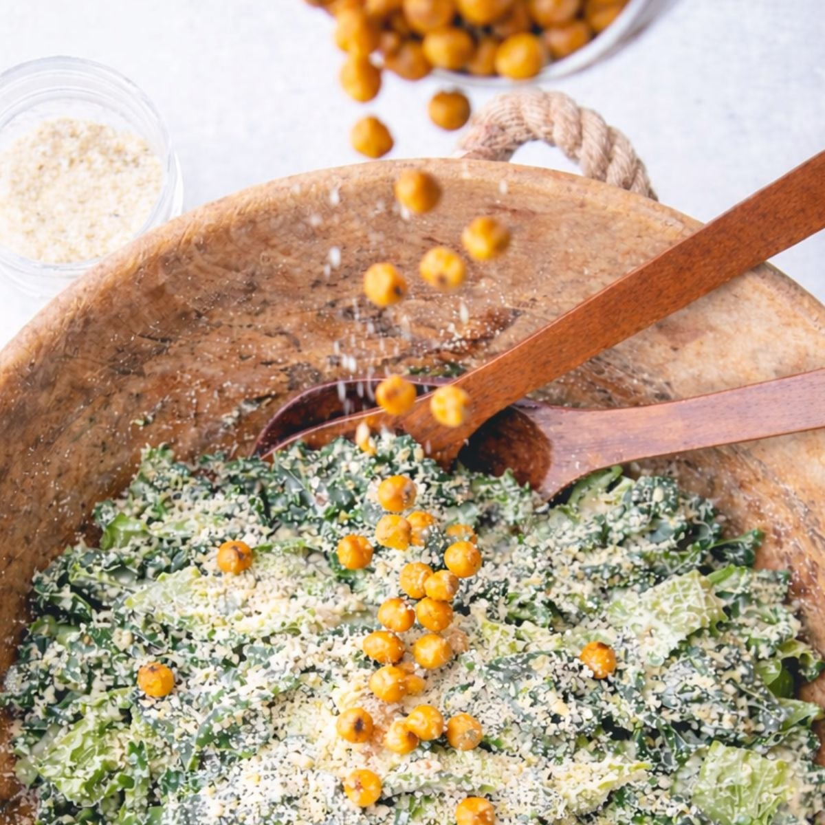 Roasted chickpea croutons and grated parmesan falling onto a bowl of dressed kale caesar salad.