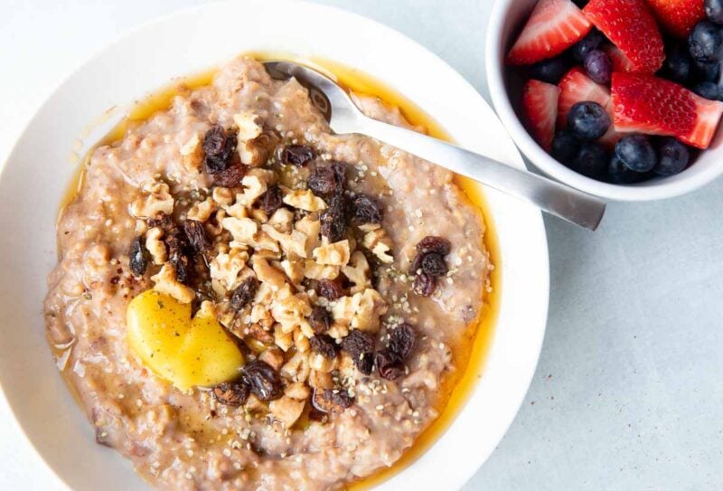 A bowl of steel cut oatmeal topped with fruit, nuts, butter, and maple syrup sits beside a small bowl of fresh berries.