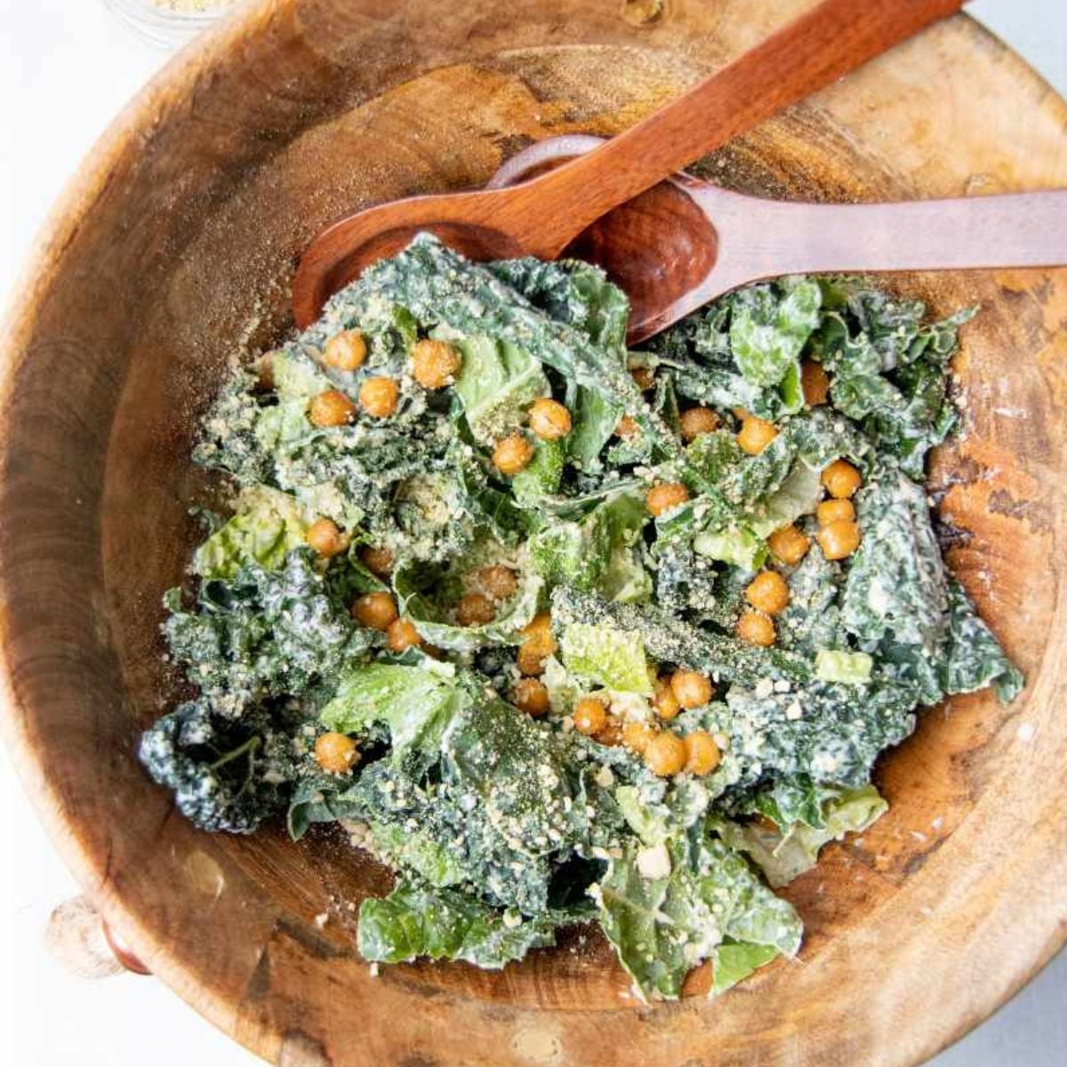Kale caesar salad tossed with chickpea croutons and grated parmesan, a wooden spoon resting across the bowl.