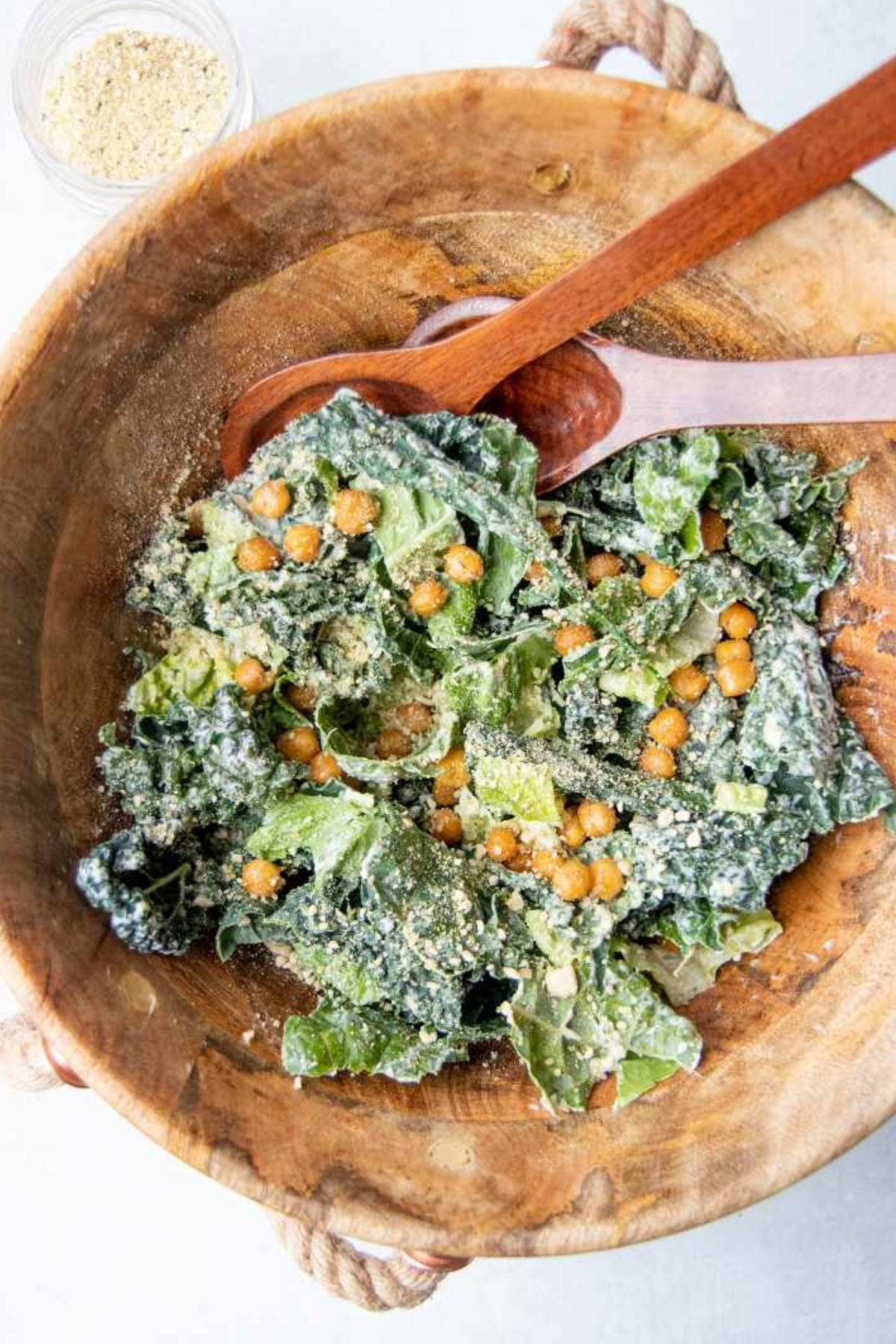 Kale caesar salad tossed with roasted chickpea croutons and grated parmesan in a wood bowl with rope handles, two wooden spoons resting across the top.