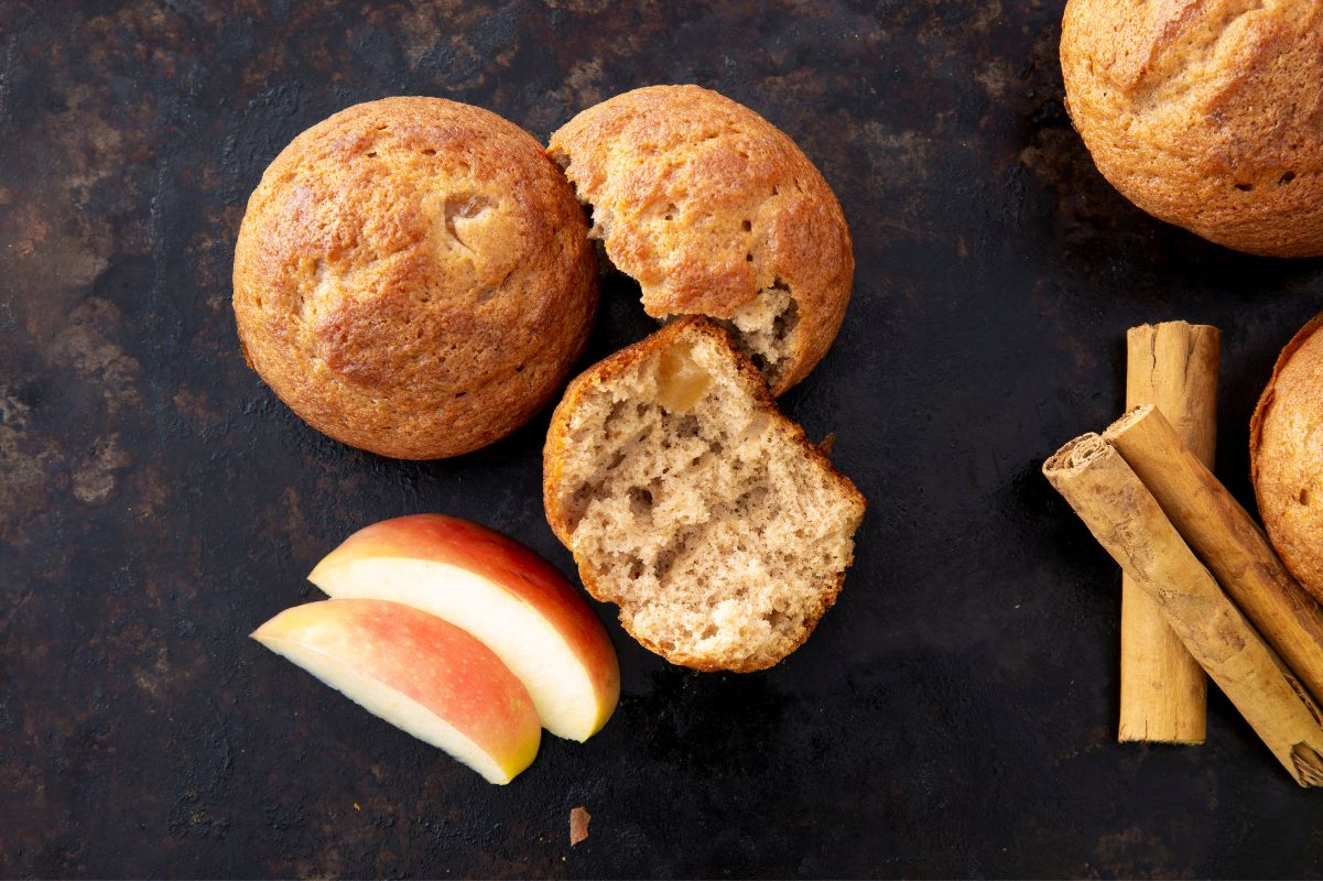 Close view of a split apple cinnamon muffin beside a whole muffin, apple slices and cinnamon sticks beside them.