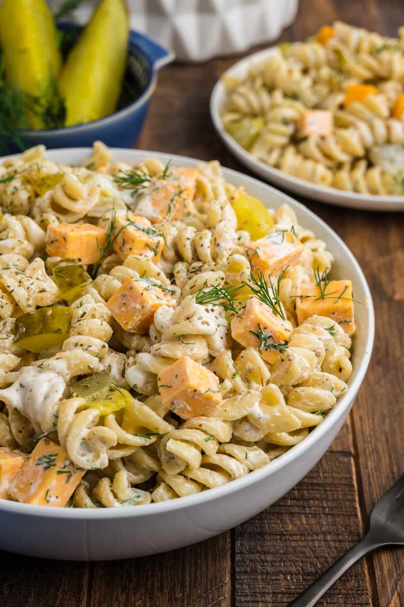 A large white serving bowl filled with dill pickle pasta salad, a bowl of dill pickle spears behind it.