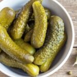 Overhead of a white bowl filled with whole dill pickles on a wooden table.