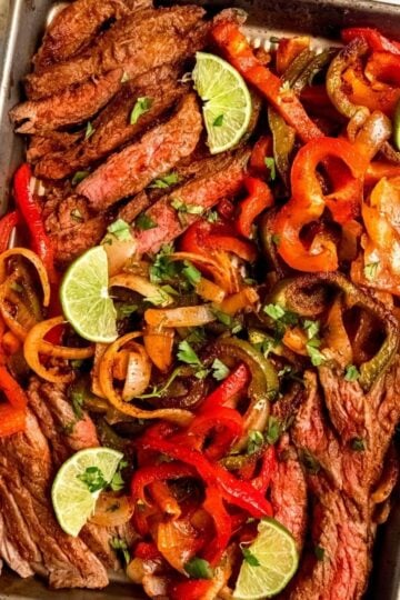 Slices of cooked steak and bell peppers cooked on a sheet pan with lime wedges